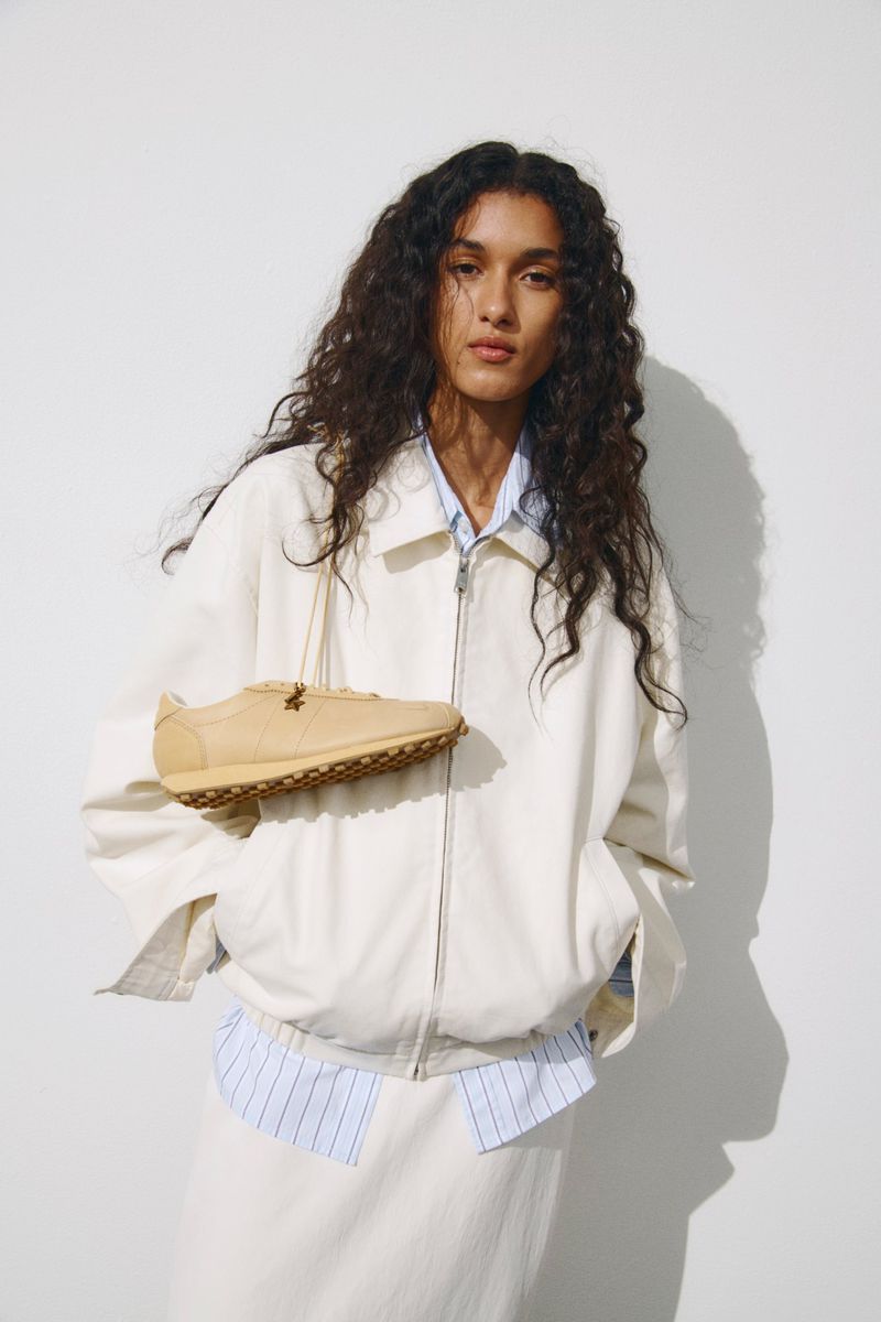 Model with long dark curly hair wearing an off-white zip-up jacket with a tan sneaker hanging around her neck against a white wall.