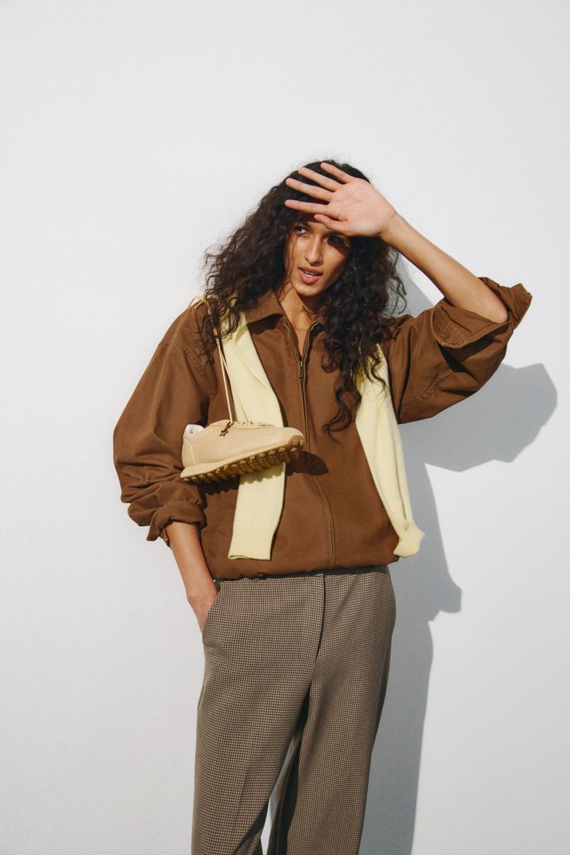 Model shielding eyes wearing a brown jacket, taupe trousers, and a draped cream knit holding a beige sneaker on a white background.