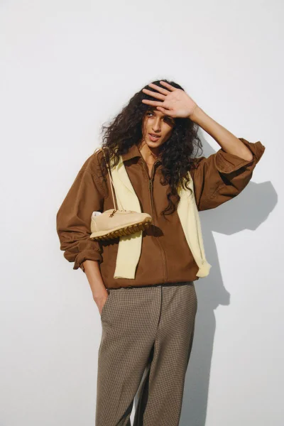 Model shielding eyes wearing a brown jacket, taupe trousers, and a draped cream knit holding a beige sneaker on a white background.