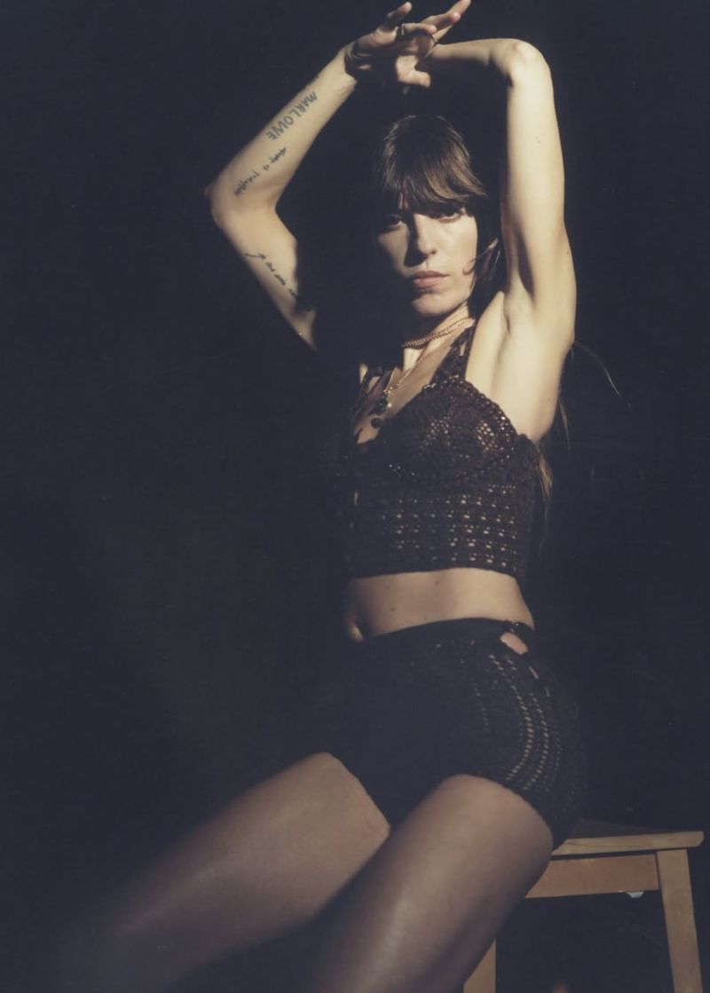 Woman in dark, shimmery crop top and shorts poses seated on a stool with arms raised against a dark background, looking at the camera.