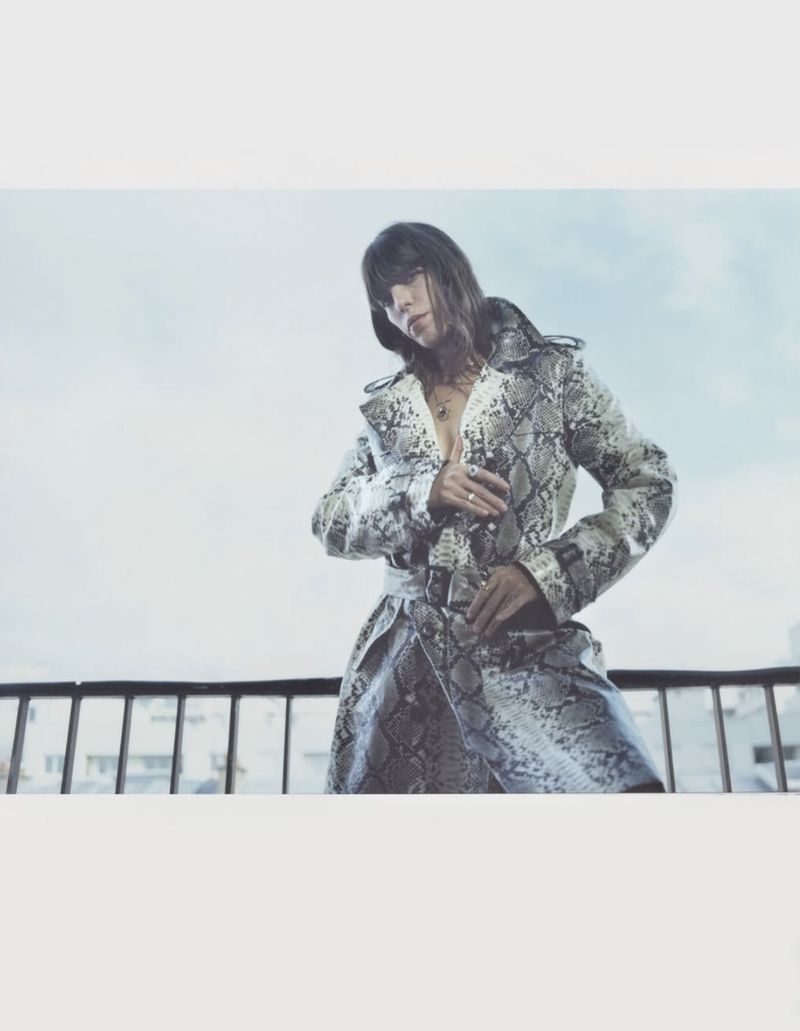 Lou Doillon wearing a snakeskin print trench coat standing on a balcony against a pale sky.