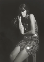 Lou Doillon seated in an embellished circular chainmail mini dress against a dark backdrop in black and white.