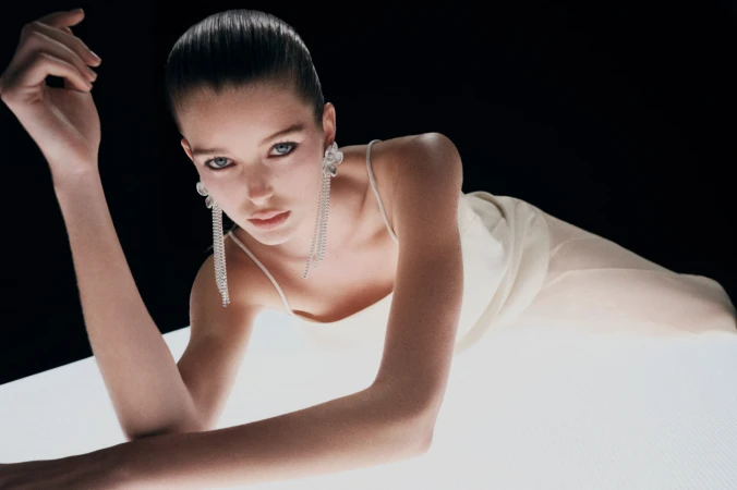 Model wearing a light dress and long crystal earrings reclining on an illuminated platform against a black background