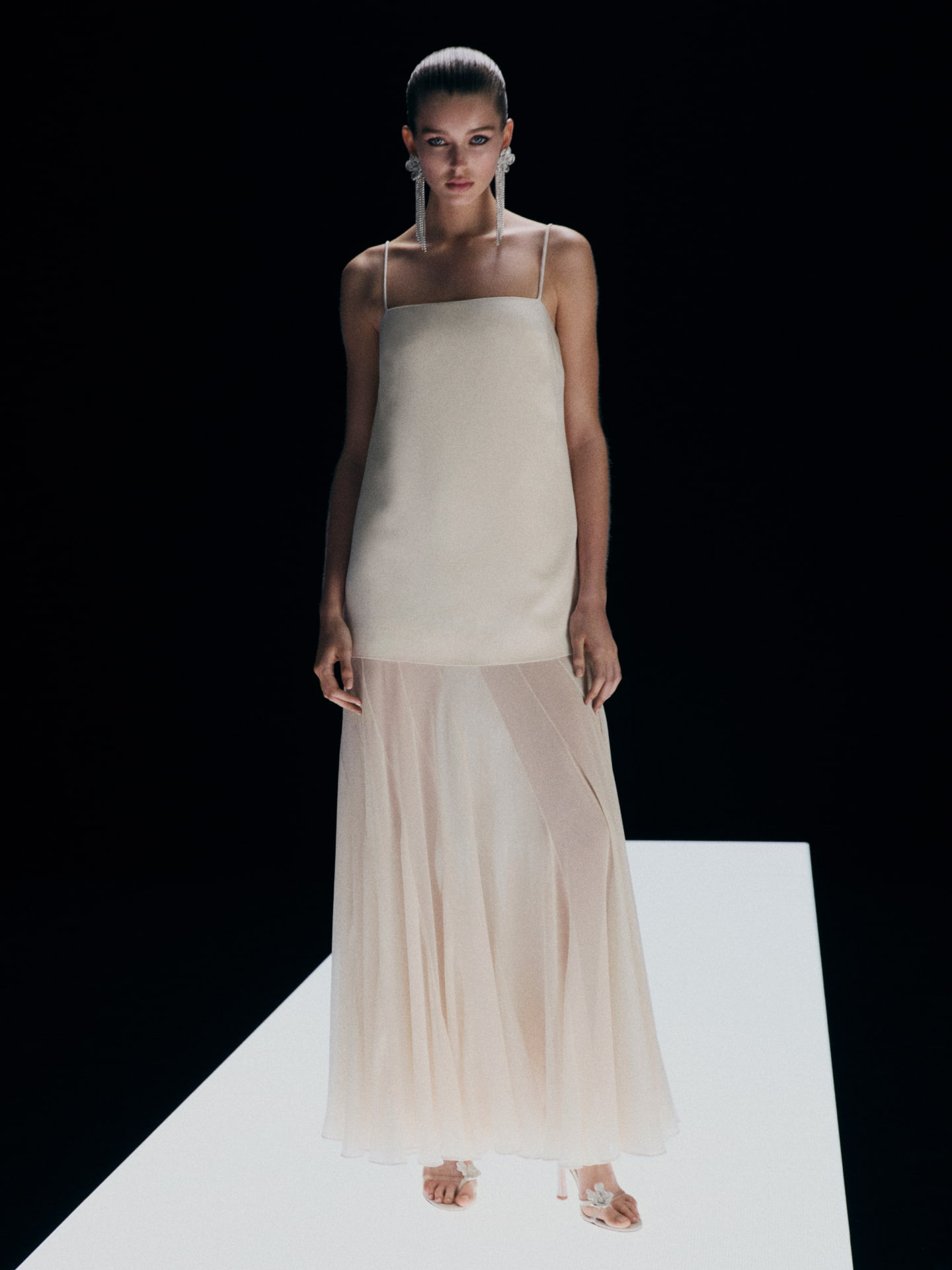 Fashion model in a full-length pose on a white platform wearing a light beige dress with spaghetti straps and a sheer skirt.