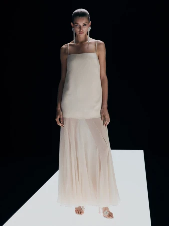 Fashion model in a full-length pose on a white platform wearing a light beige dress with spaghetti straps and a sheer skirt.