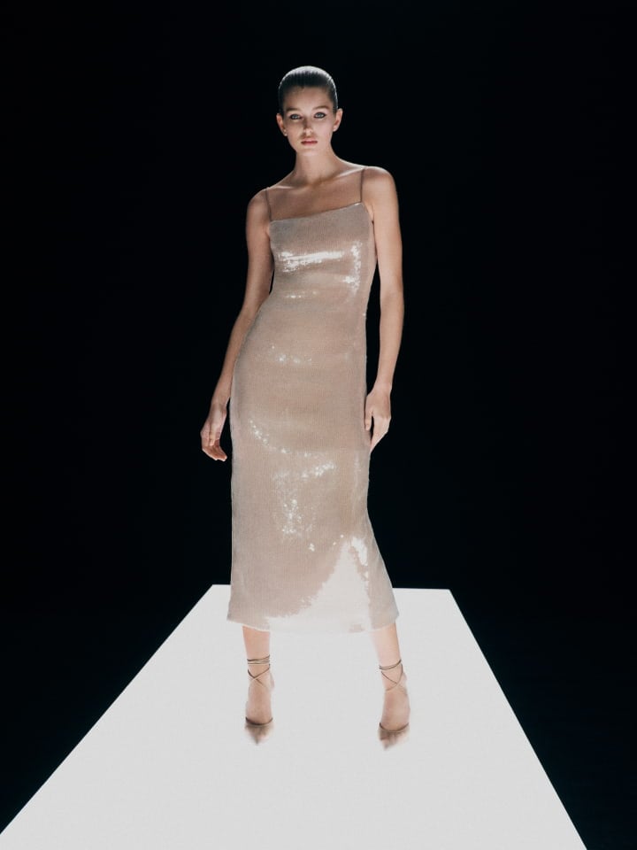 Elegant woman in shimmering midi dress standing on a white platform against a black background in a fashion forward style.
