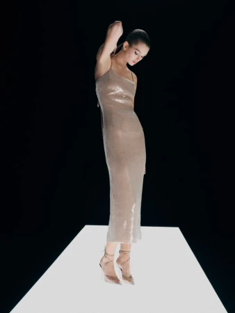 A slender woman in a champagne dress poses against a black backdrop. She stands on a white surface with minimal background distractions.