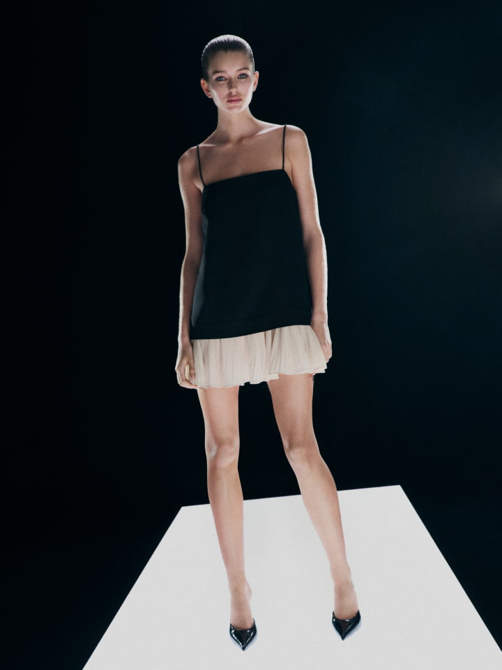 Model standing on a lighted platform wearing a black camisole top with a pleated beige mini skirt against a dark background