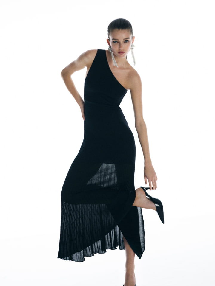 Model in a black one-shoulder dress and high heels posing against a bright white background