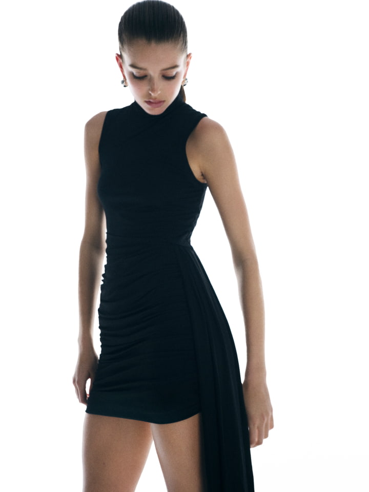 Fashion model poses in a sleeveless, black, mock turtleneck mini dress with draped side detail against a bright, gradient white backdrop.