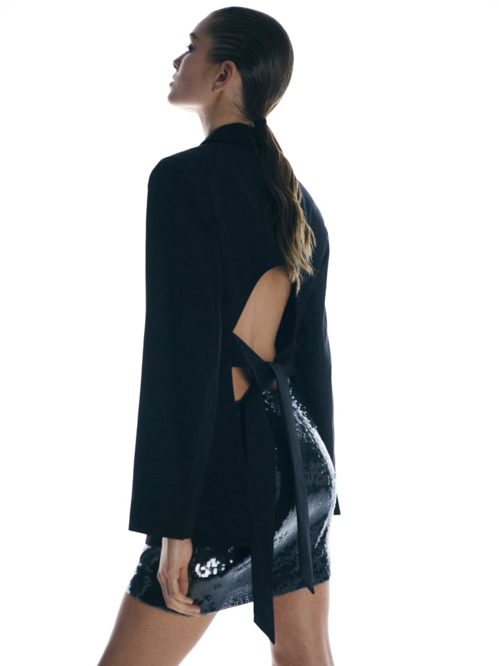 Model wearing a black open-back blazer and sequin skirt against a bright white background