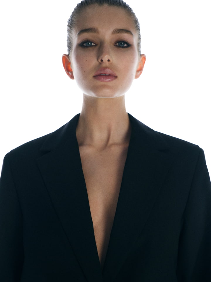 Headshot of a blonde adult woman with fair skin, wearing a black blazer, looking at the camera against a bright white backdrop.