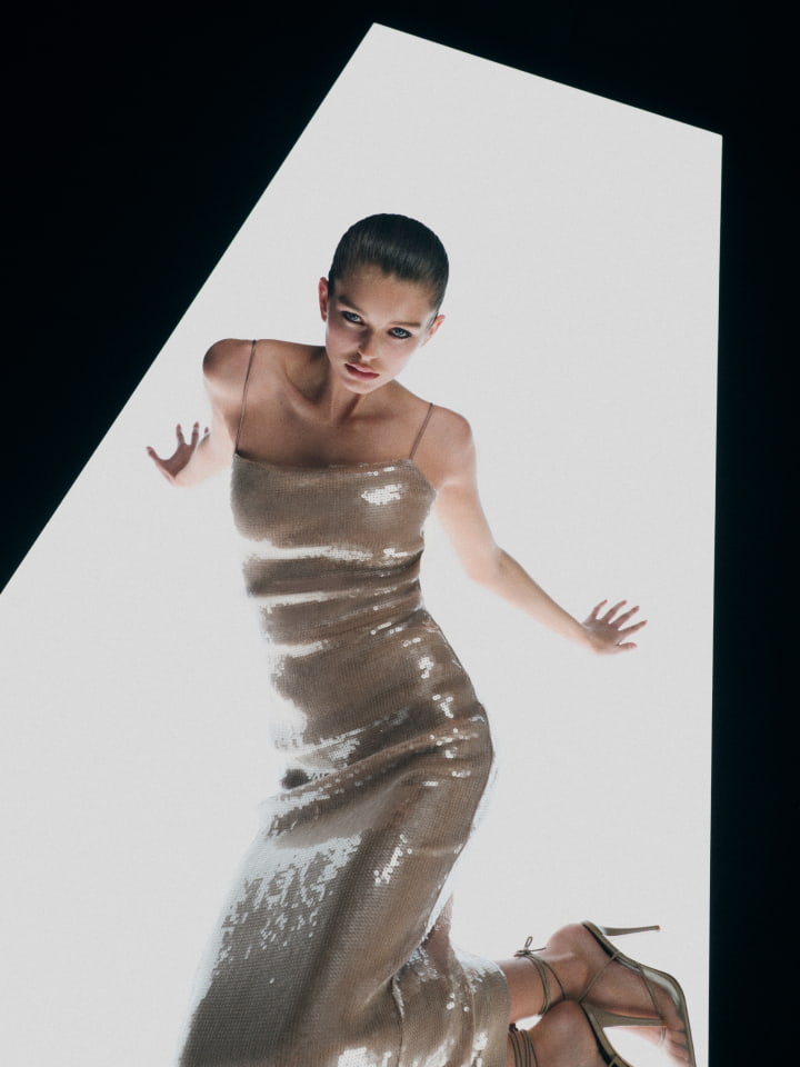 Woman in shimmering champagne-colored gown with spaghetti straps poses dynamically on a white platform against a black backdrop.
