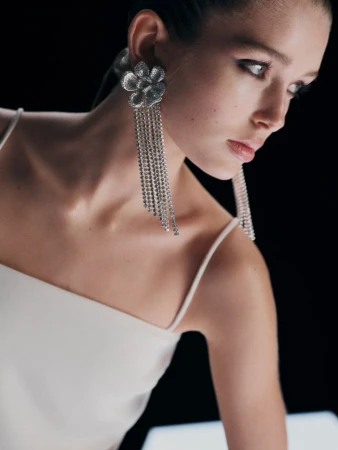 Close-up of a model wearing a white top and elaborate crystal fringe flower earrings against a black background
