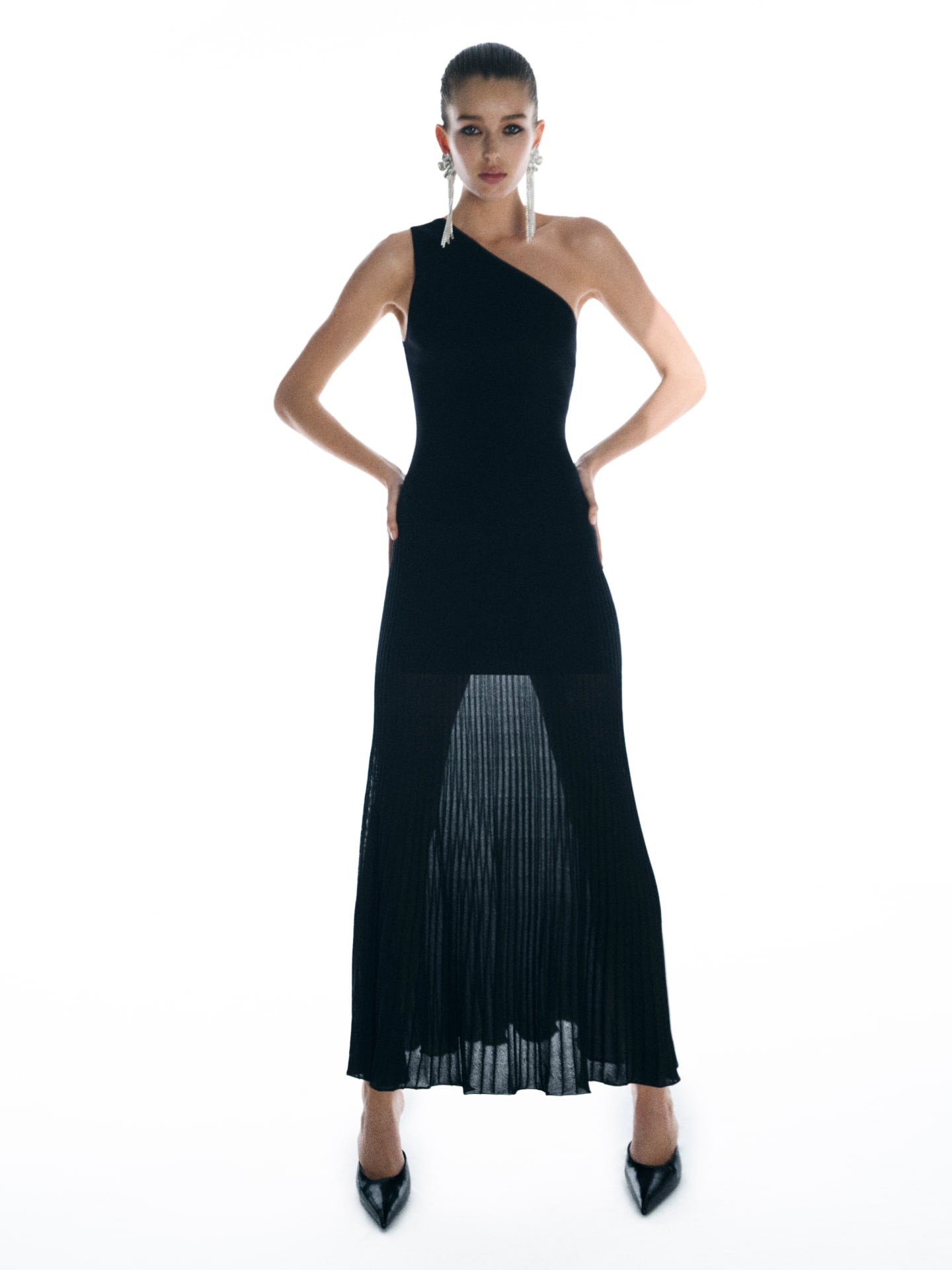 Model in a black one-shoulder maxi dress and statement earrings standing against a bright white background