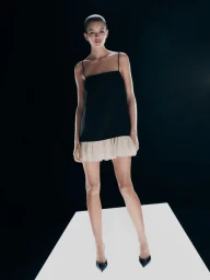 Model standing on a lighted platform wearing a black camisole top with a pleated beige mini skirt against a dark background