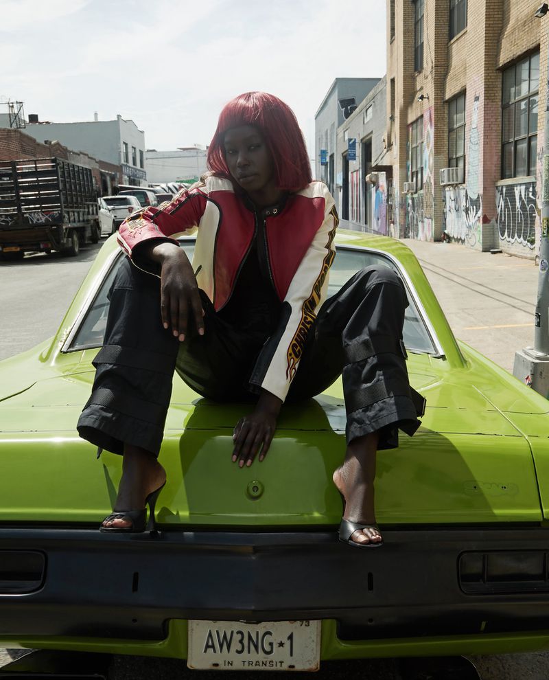 A Black woman with red hair poses atop a lime-green car in a red-and-white racing jacket and black pants on an urban street.