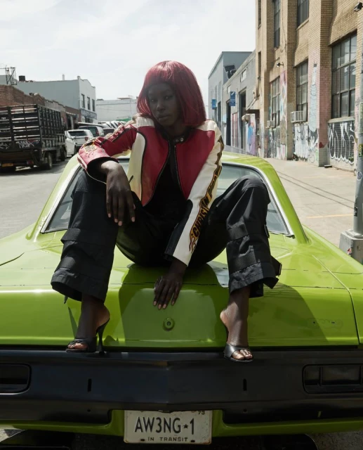 A Black woman with red hair poses atop a lime-green car in a red-and-white racing jacket and black pants on an urban street.