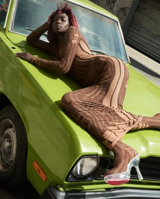 A Black female fashion model in a brown patterned dress and clear platform shoes poses on a green vintage car, outdoor urban setting.