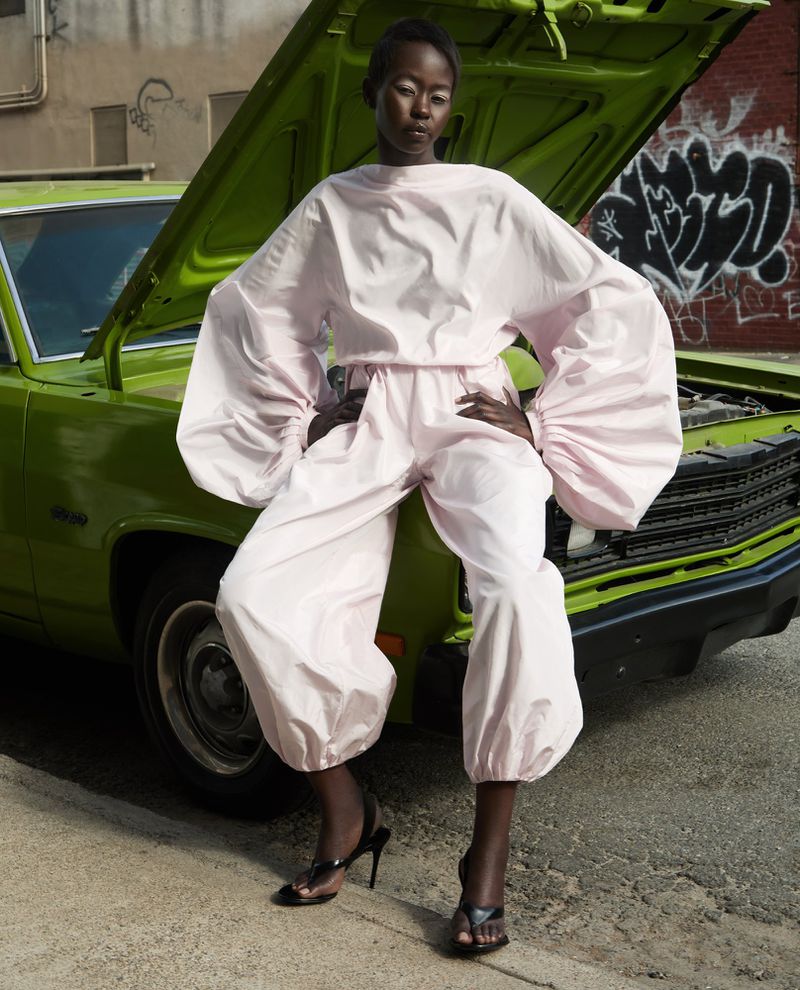 Fashion editorial shot of a Black woman in a light pink outfit posing on the hood of a green car in front of a brick wall.