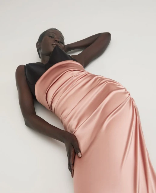 Elegant woman poses in a striking black and pink satin dress, lying against a neutral backdrop in a fashion-forward portrait.