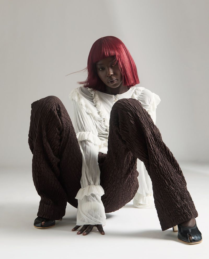 Dark-skinned woman with red bob, wearing a white frilly shirt and brown textured pants, sits in a studio on a white background.