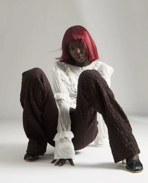 Dark-skinned woman with red bob, wearing a white frilly shirt and brown textured pants, sits in a studio on a white background.