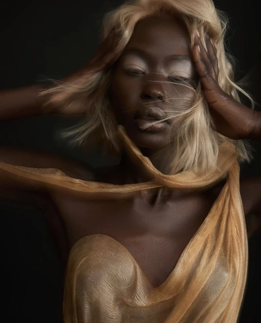 Striking portrait of an African woman with blonde hair and hands framing her face, draped in a gold fabric, against a dark background.