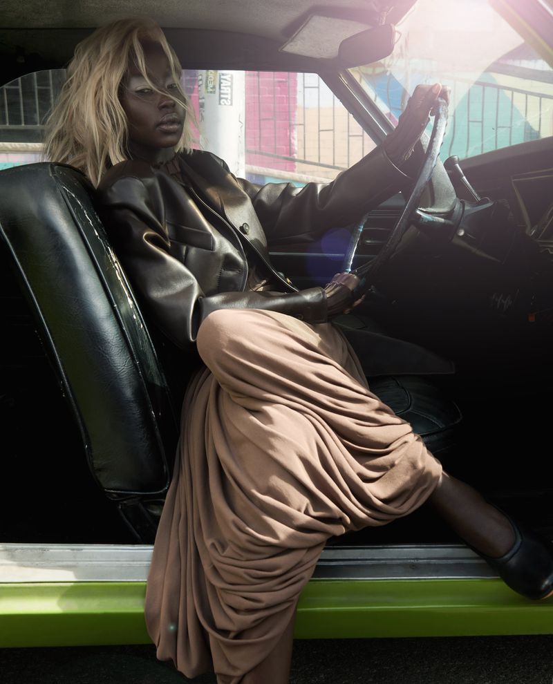 Model Aweng Chuol sits in a vintage car wearing a dark leather jacket and draped beige skirt for Numéro Netherlands.
