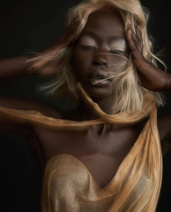 Aweng Chuol with blonde hair wearing a gold sheer garment with hands framing face in Numéro Netherlands editorial.