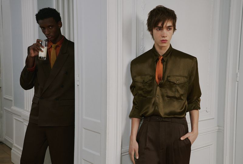 Two young male models in tailored suits posing in an interior hallway with white panelled walls.