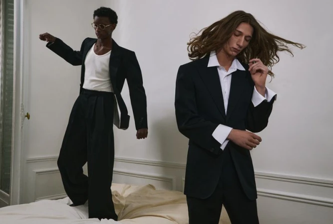 Two male models in tailored black suits and white shirts, one standing in foreground and one posing on a bed.