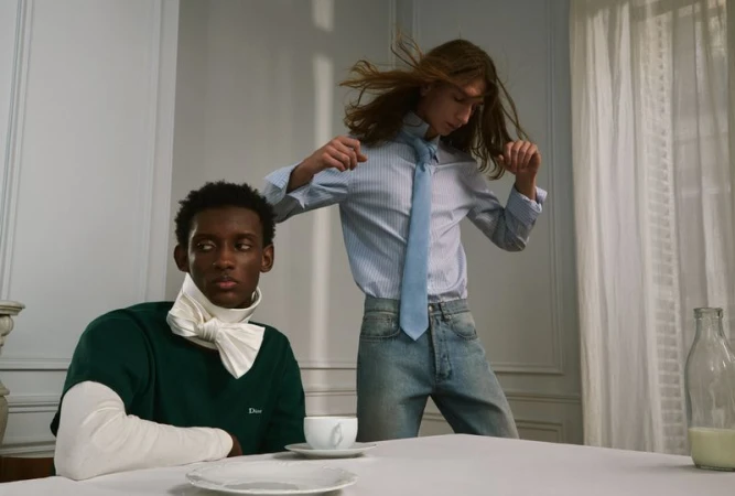 Two male models in tailored shirts and denim posing at a table with a white tablecloth in an interior editorial scene.