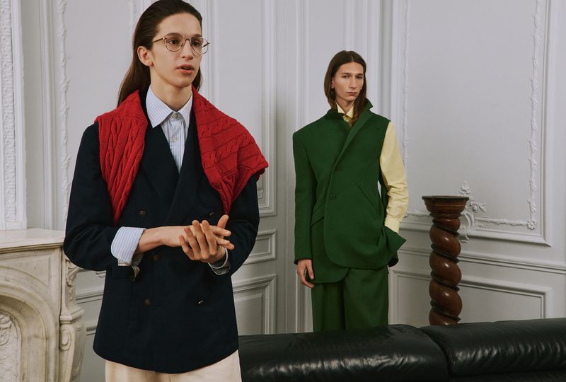 Two models in tailored menswear and layered knitwear posing in an interior room with white wall mouldings and a black leather couch.