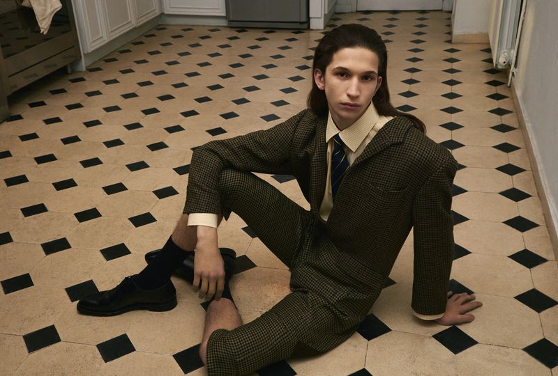 Man in a tailored check short suit and tie seated on a black and cream tiled kitchen floor.