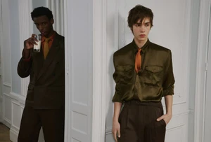 Two young male models in tailored suits posing in an interior hallway with white panelled walls.