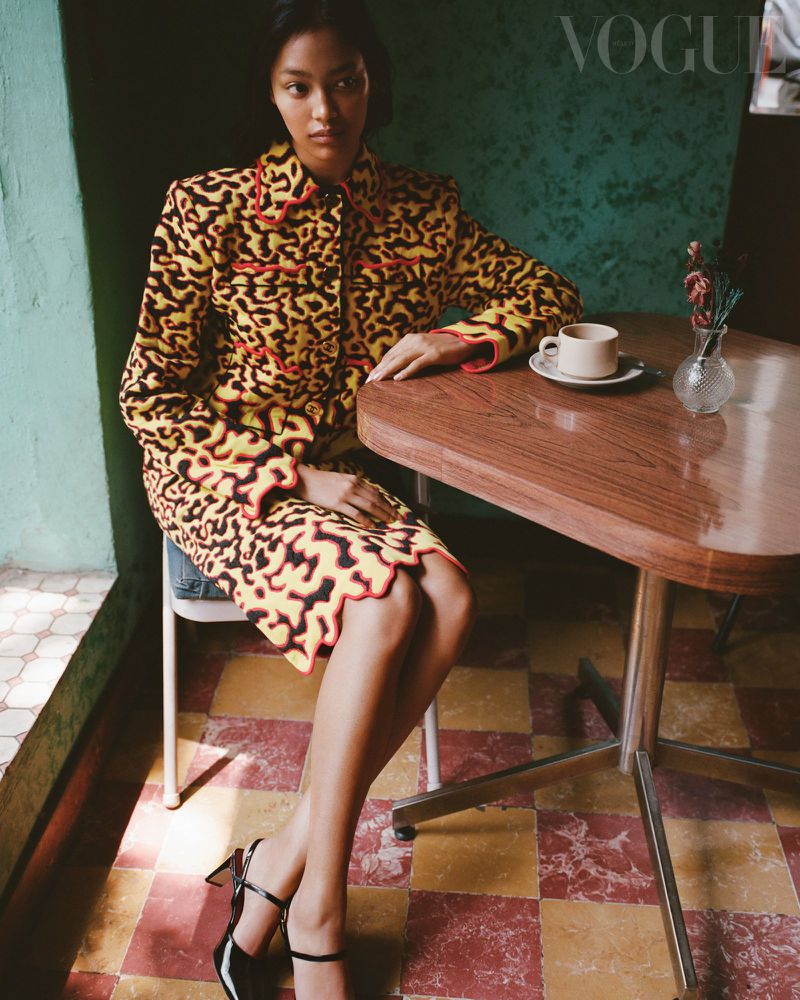 Model Aneken Zaldivar in a yellow and black patterned skirt suit seated beside a wooden table in a cafe.