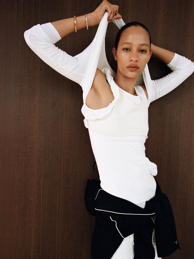 Model Selena Forrest pulling a white long-sleeve top over her head against a dark wood paneled backdrop.