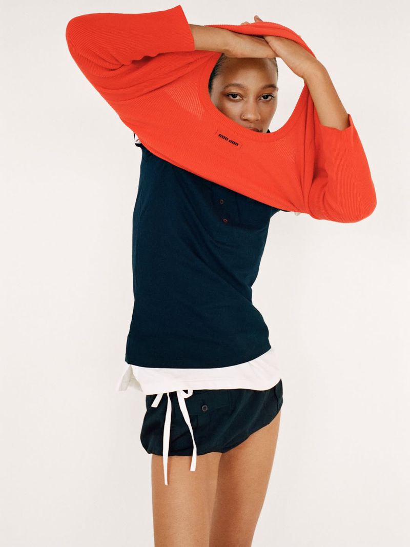 Model Selena Forrest pulling a bright red crewneck sweater over her head against a plain white studio background.