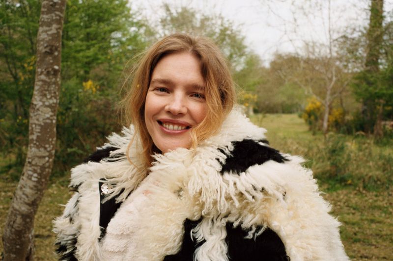 Natalia Vodianova smiling outdoors in a fluffy black and white shearling coat against a natural woodland background.