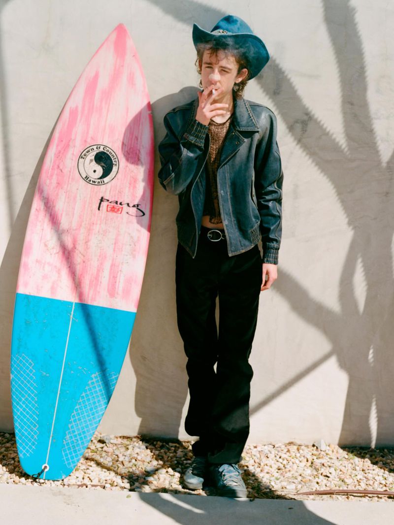 A man in a blue cowboy hat and leather jacket smoking while leaning against a wall next to a pink and blue surfboard.
