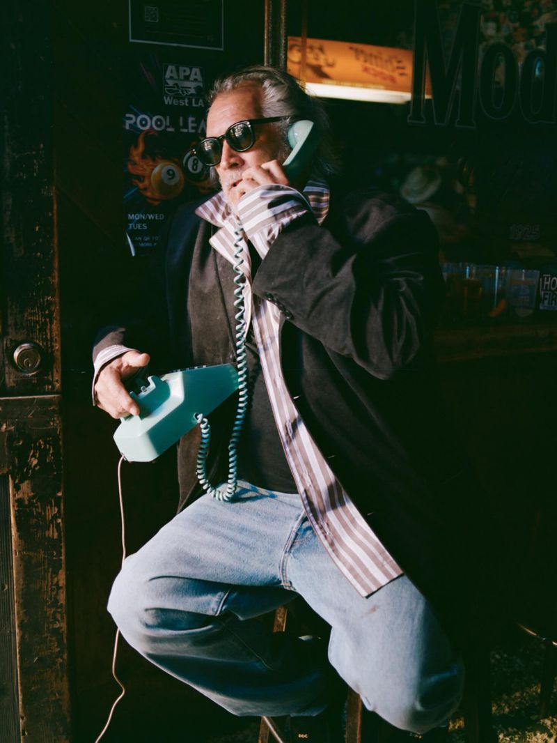 Man with grey hair in sunglasses and black jacket talking on a vintage teal corded phone while seated in a doorway.