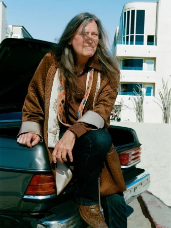 An older man with long hair wearing a patterned brown coat and jeans sits on the trunk of a vintage car outside a beach house.