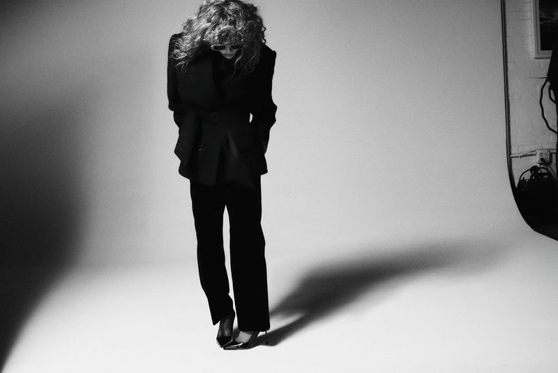 Natasha Lyonne standing in a black tailored suit and pointed heels against a white studio seamless backdrop with a dramatic shadow.