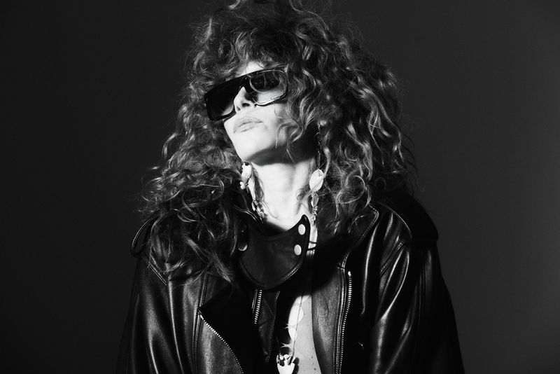Natasha Lyonne in oversized sunglasses and a black leather jacket with voluminous curly hair in a black and white portrait.