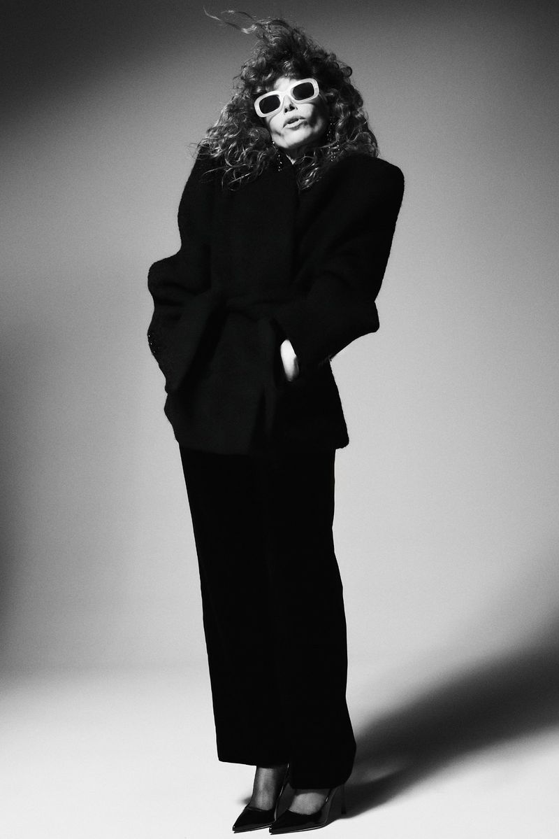 Natasha Lyonne wearing white sunglasses and a black coat against a grey gradient background in a black and white portrait.