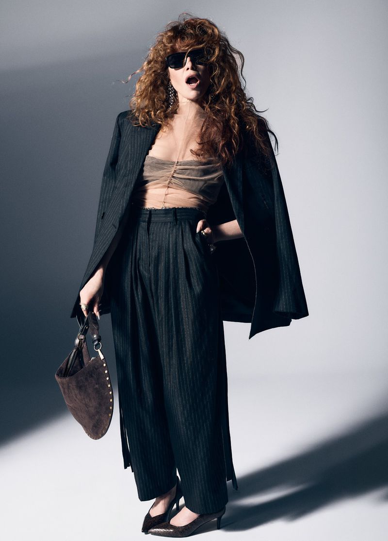 Natasha Lyonne wearing a dark pinstripe suit, sunglasses, and sheer top while holding a brown suede bag against a studio backdrop.
