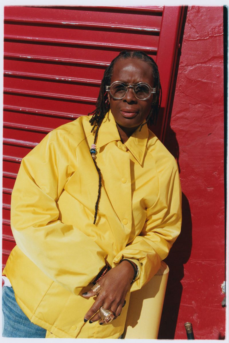 Mickalene Thomas wearing a bright yellow jacket and round clear eyeglasses while leaning against a vibrant red corrugated wall.