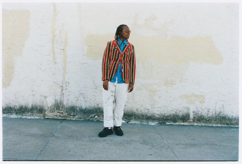 Mickalene Thomas posing on a sidewalk against a textured off-white wall in a red striped blazer and white paint-splattered pants.