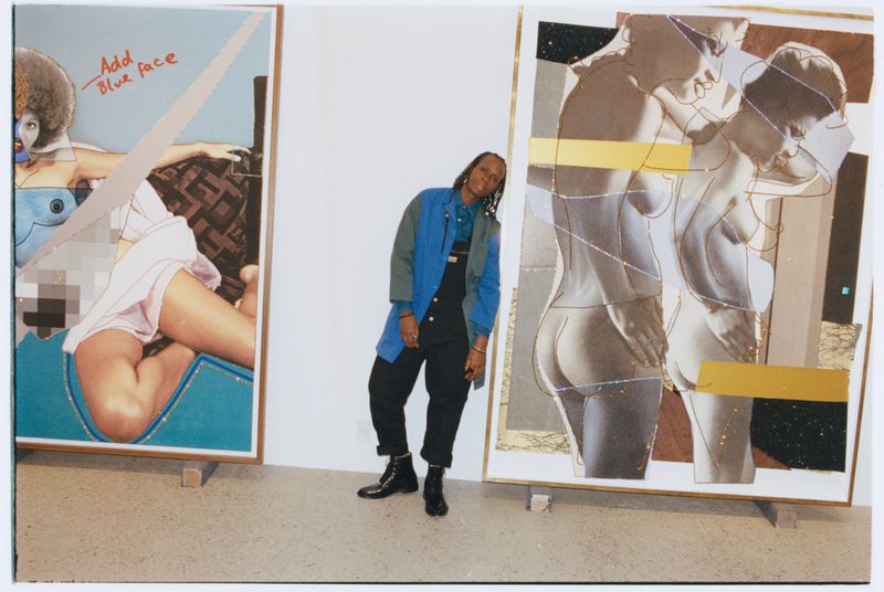 Person standing between two large mixed-media art canvases in a gallery space wearing a blue jacket and dark overalls.